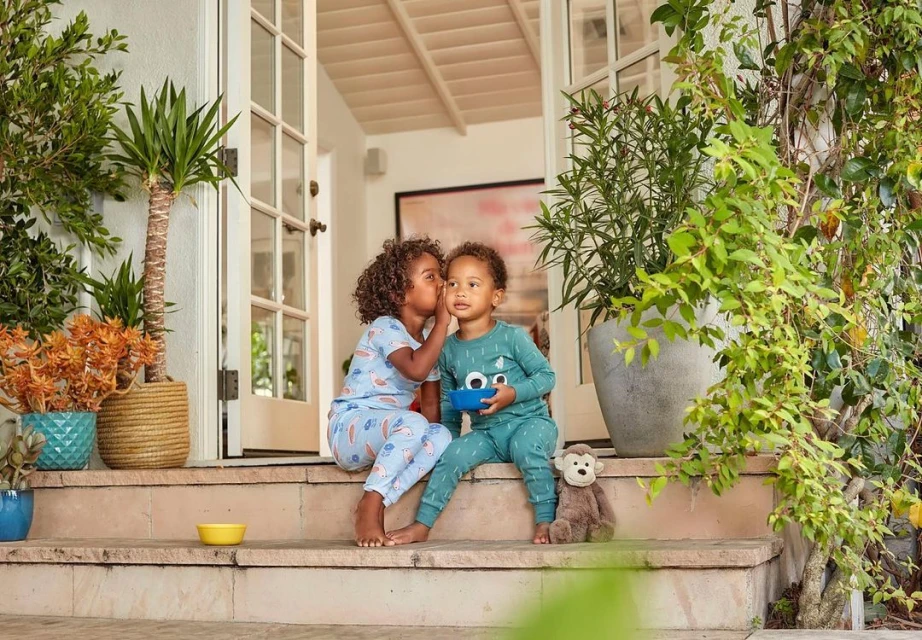 Two children in pajamas sit on steps outside a house with potted plants. One child whispers in the other's ear in a candid moment.