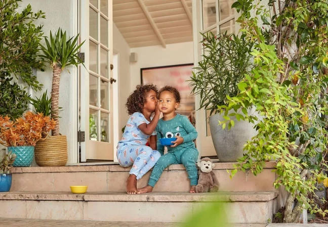 Two children in pajamas sit on steps outside a house with potted plants. One child whispers in the other's ear in a candid moment.