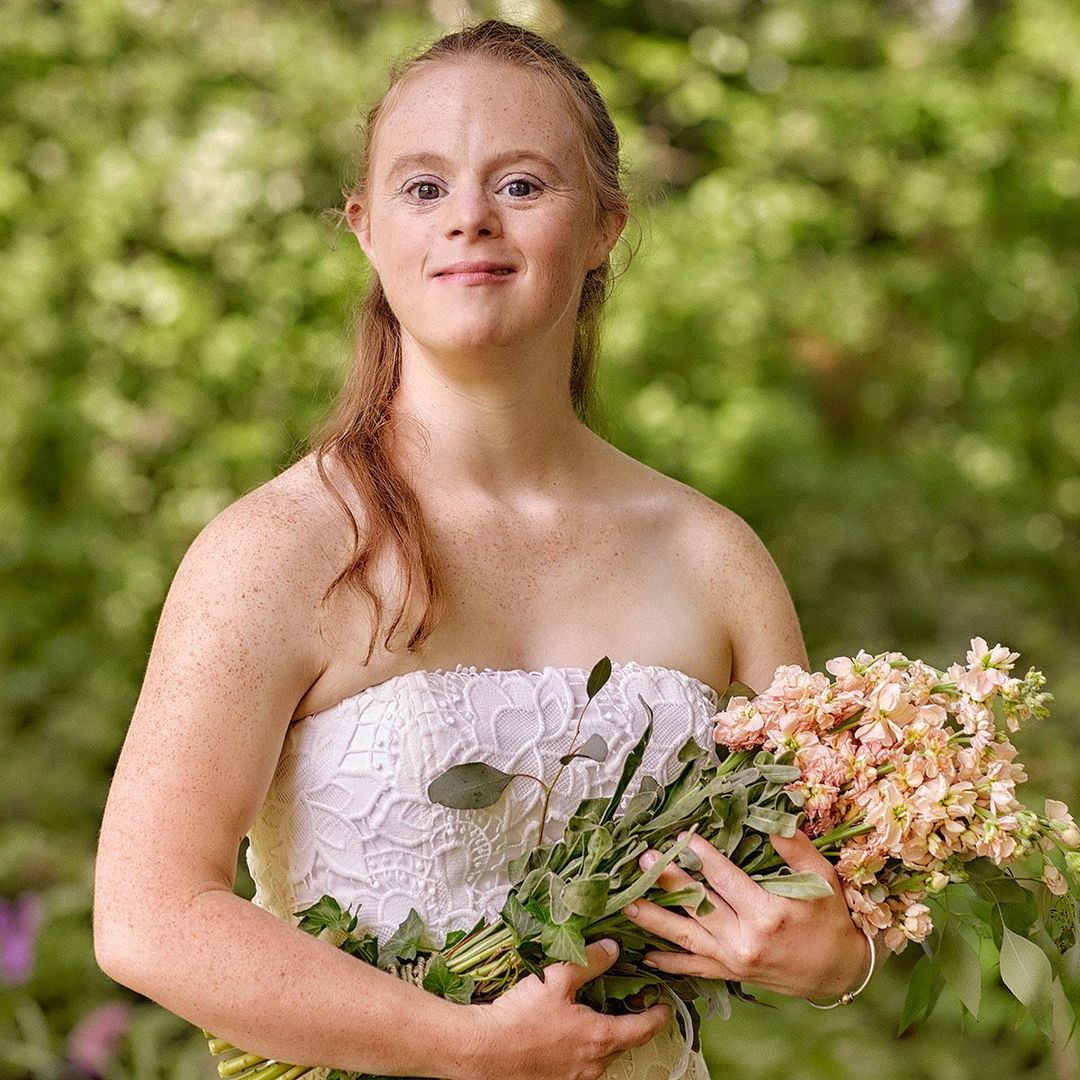 Portrait of a woman with Down syndrome in a strapless white dress, holding a bouquet of flowers, with a blurred leafy green background.