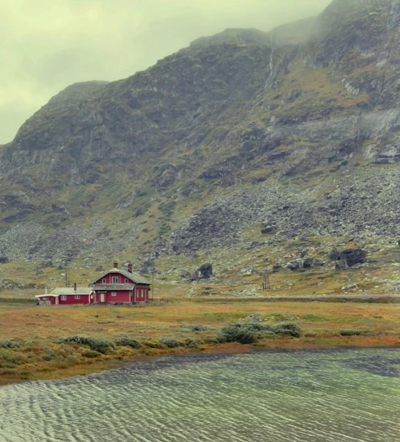 Scenic view of a red cabin house by a lake, set against a backdrop of a vast, rocky mountain under an overcast sky, creating a tranquil scene.