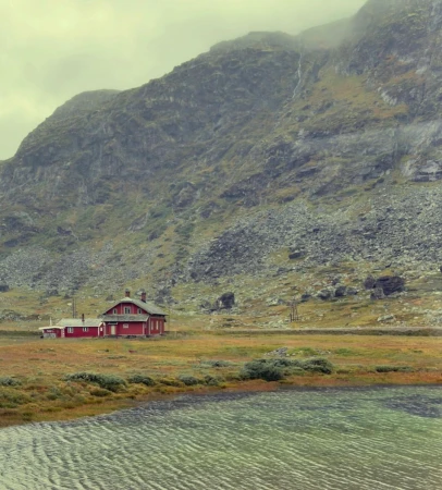 Scenic view of a red cabin house by a lake, set against a backdrop of a vast, rocky mountain under an overcast sky, creating a tranquil scene.