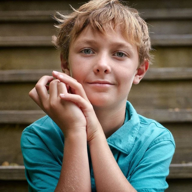 Portrait of a young boy with blond hair and blue eyes sitting on wooden steps, looking directly at the camera with a gentle smile.