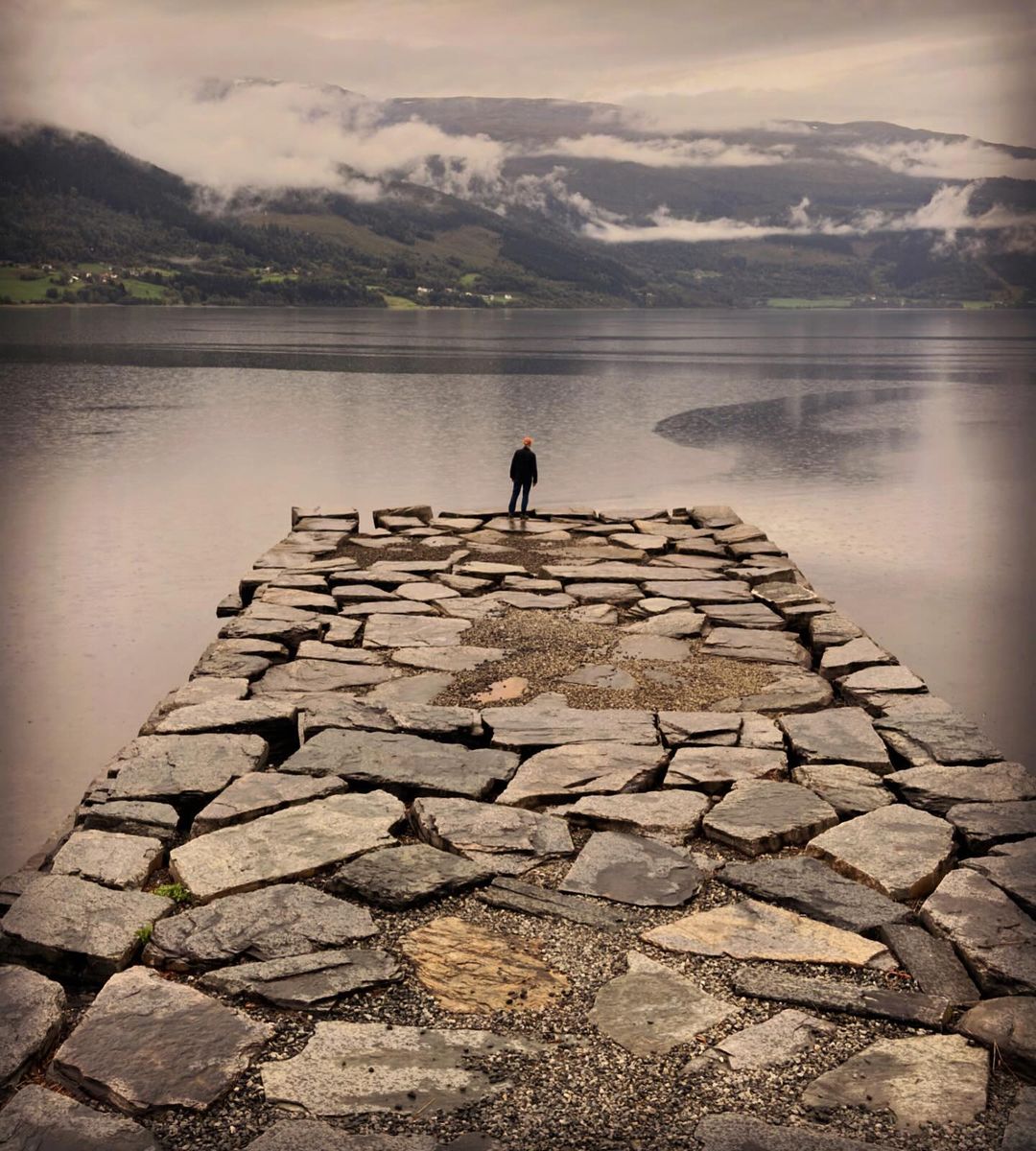 Person standing on a stone pier overlooking a large lake surrounded by mountains shrouded in mist and clouds on an overcast day.