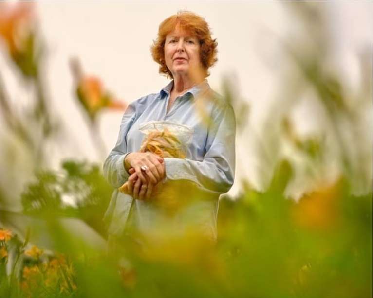 Adult woman with red hair stands in a field with orange flowers holding a bag, creating a warm and natural outdoor portrait.