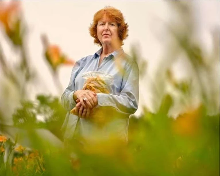 Adult woman with red hair stands in a field with orange flowers holding a bag, creating a warm and natural outdoor portrait.