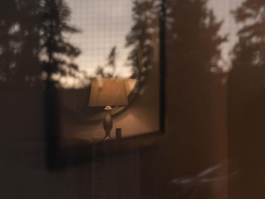 Illuminated lamp on bedside table inside a cozy room, viewed through a glass panel with trees visible outside the window.
