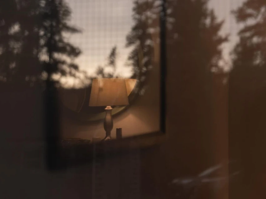 Illuminated lamp on bedside table inside a cozy room, viewed through a glass panel with trees visible outside the window.