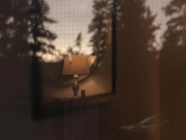 Illuminated lamp on bedside table inside a cozy room, viewed through a glass panel with trees visible outside the window.