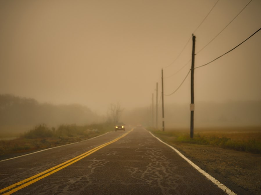 Car driving on a rural road through dense fog with power lines in the background, creating a mysterious and atmospheric scene.