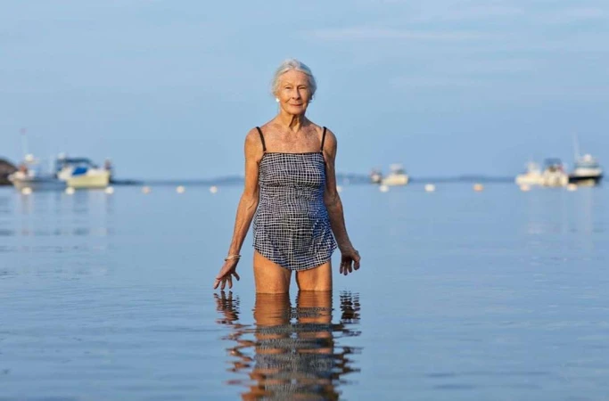 Elderly woman standing waist-deep in calm ocean water wearing a patterned swimsuit with small boats visible on the horizon.