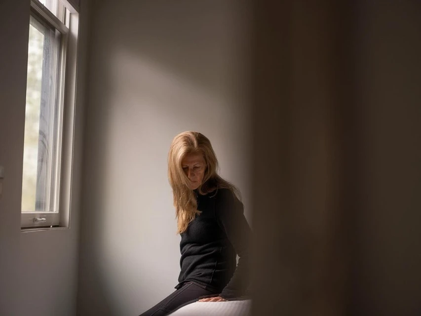 Seated woman in dark clothes gazes down in a light-filled room with a window, casting shadows on the wall and her blonde hair.