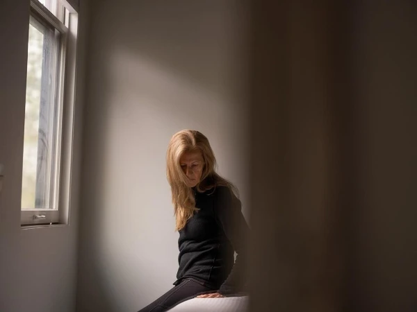 Seated woman in dark clothes gazes down in a light-filled room with a window, casting shadows on the wall and her blonde hair.