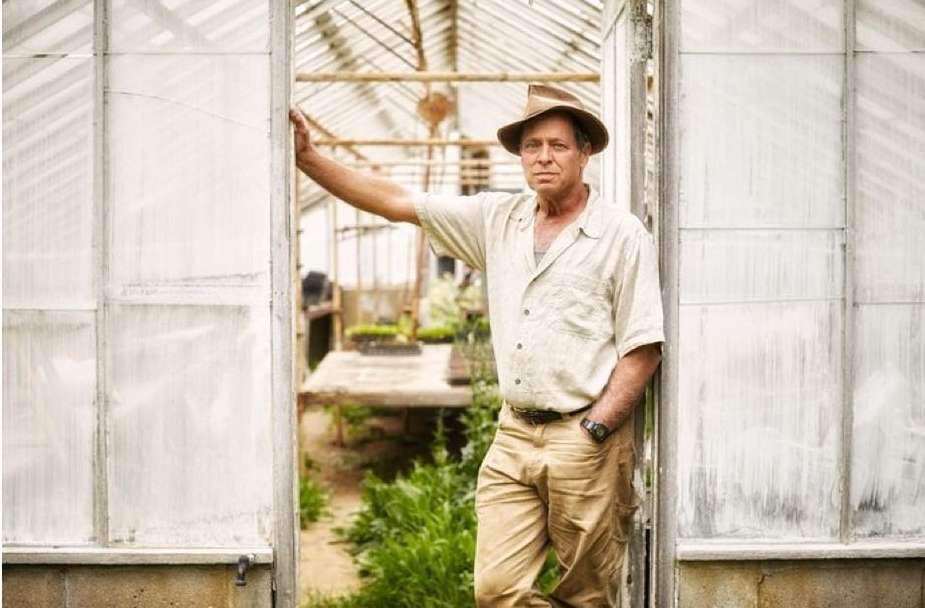 Adult male in a hat stands at the doorway of a greenhouse with plants, wearing a button-down shirt and khaki pants in daytime.
