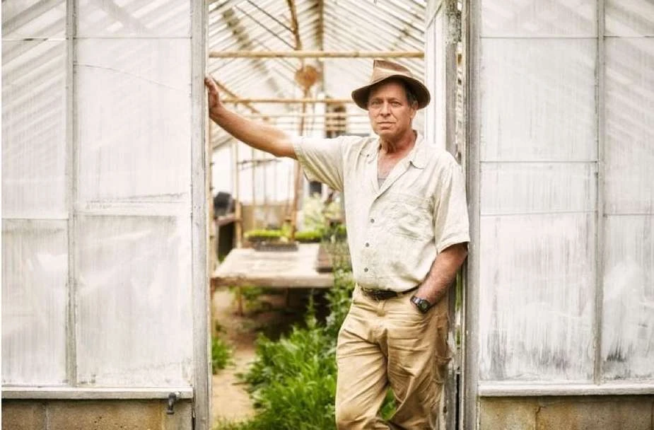 Adult male in a hat stands at the doorway of a greenhouse with plants, wearing a button-down shirt and khaki pants in daytime.
