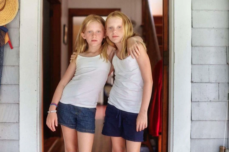 Two fair-skinned tween girls with blonde hair pose indoors wearing white tank tops and shorts, in front of a doorway with gray siding.