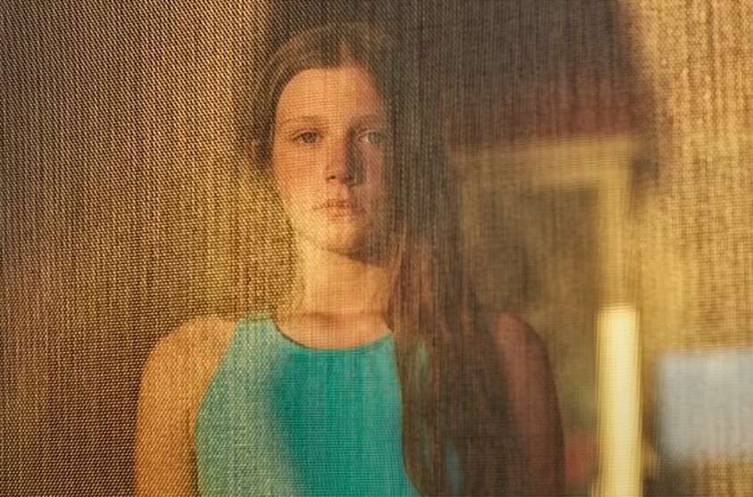 Portrait of a young woman with long brown hair, wearing a turquoise top, standing indoors with natural sunlight creating a warm mood.
