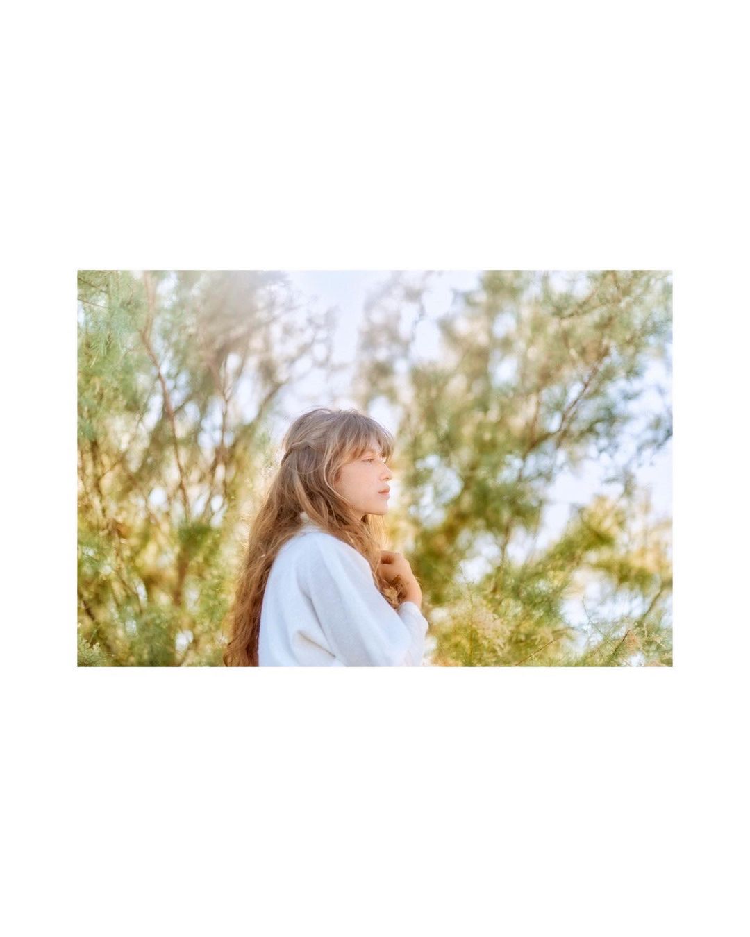 Portrait of a young woman with long brown hair, wearing a white shirt, posing outdoors with a serene expression and blurred green foliage.
