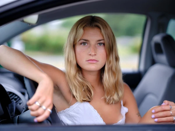 Young blond woman with freckles seated inside a car wearing a white lace top