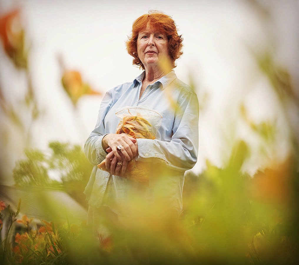 Portrait of a mature woman with red hair holding a bowl of orange lilies in a garden setting, wearing a blue button-down shirt.