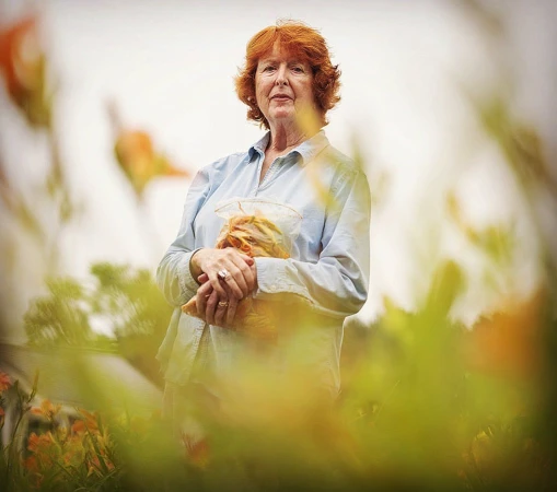 An elderly woman with red hair wearing a light blue shirt holds a bag while standing outdoors surrounded by out-of-focus flowers.