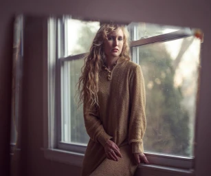 Woman with long curly blonde hair wearing a textured tan dress leaning against a window frame.