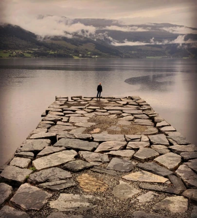 Lone figure standing at the edge of a rough stone pier overlooking a calm lake and misty mountain landscape