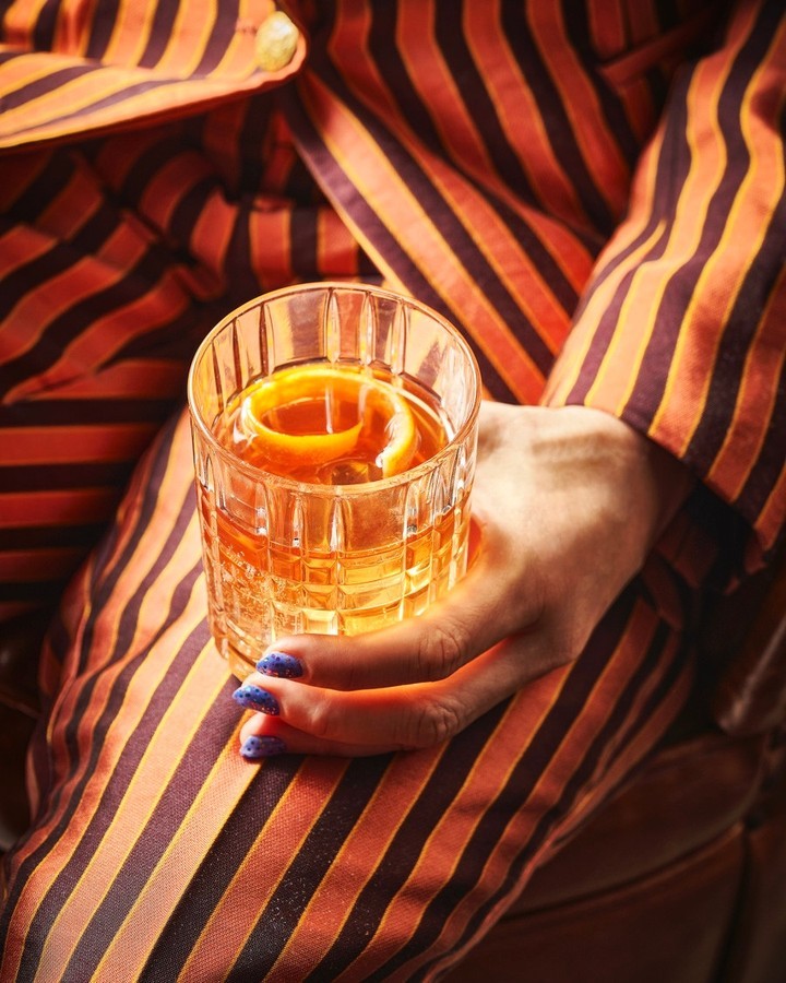 Woman holds glass of cocktail with orange peel garnish, wearing orange and brown striped pajamas; close-up, indoor setting, drink photography.