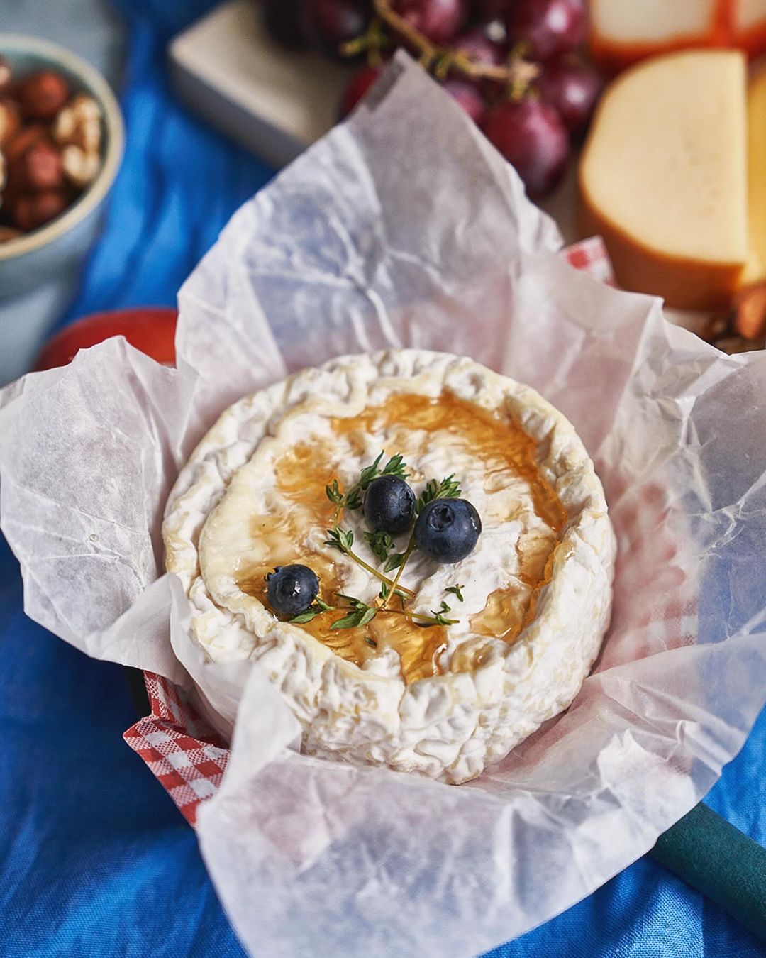 Overhead close-up of a round cheese topped with blueberries and herbs, presented in paper with grapes and nuts visible in the background.