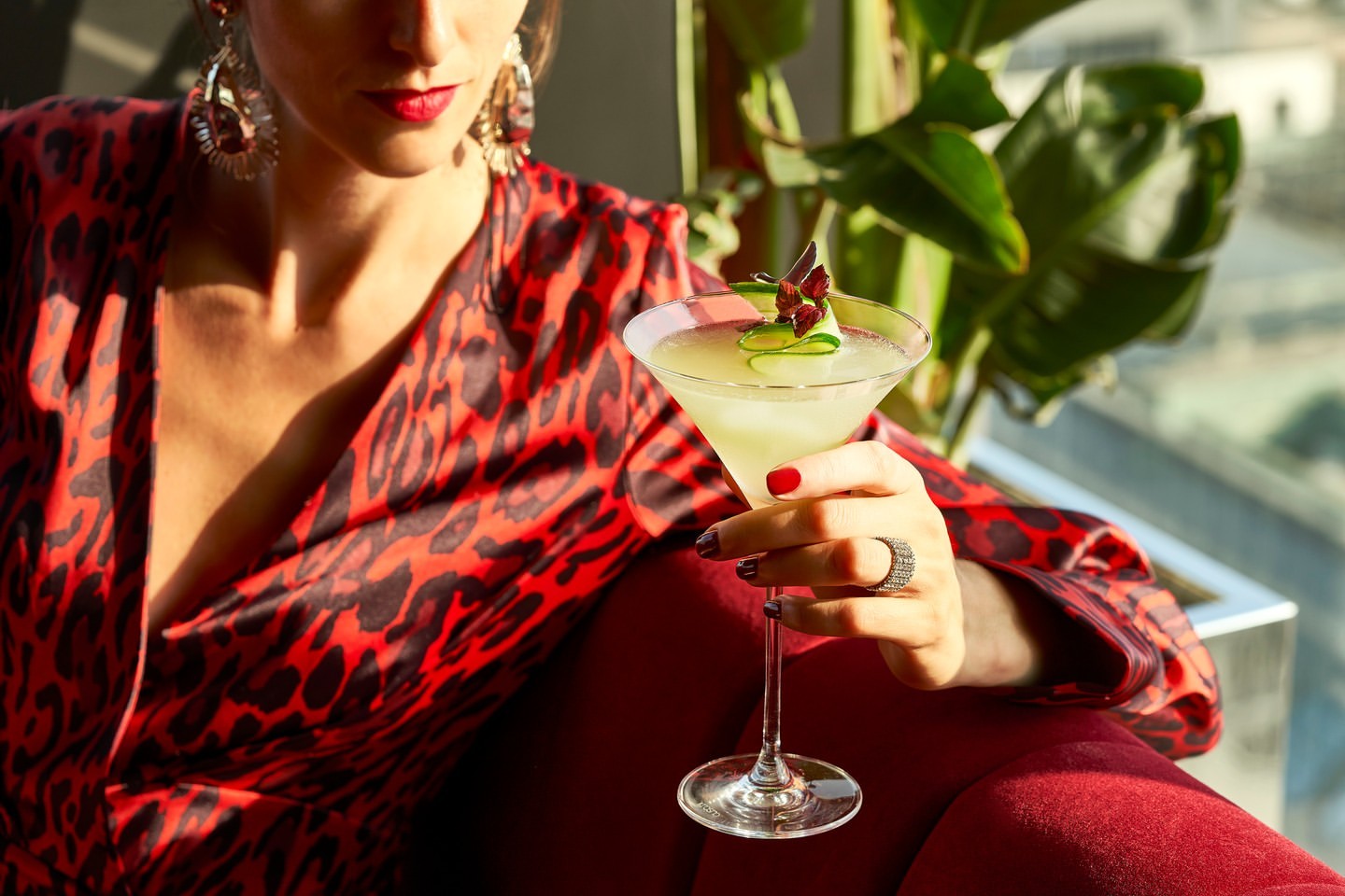 Elegant woman in animal print dress poses indoors, holding a cocktail with cucumber garnish near a window and green plant.