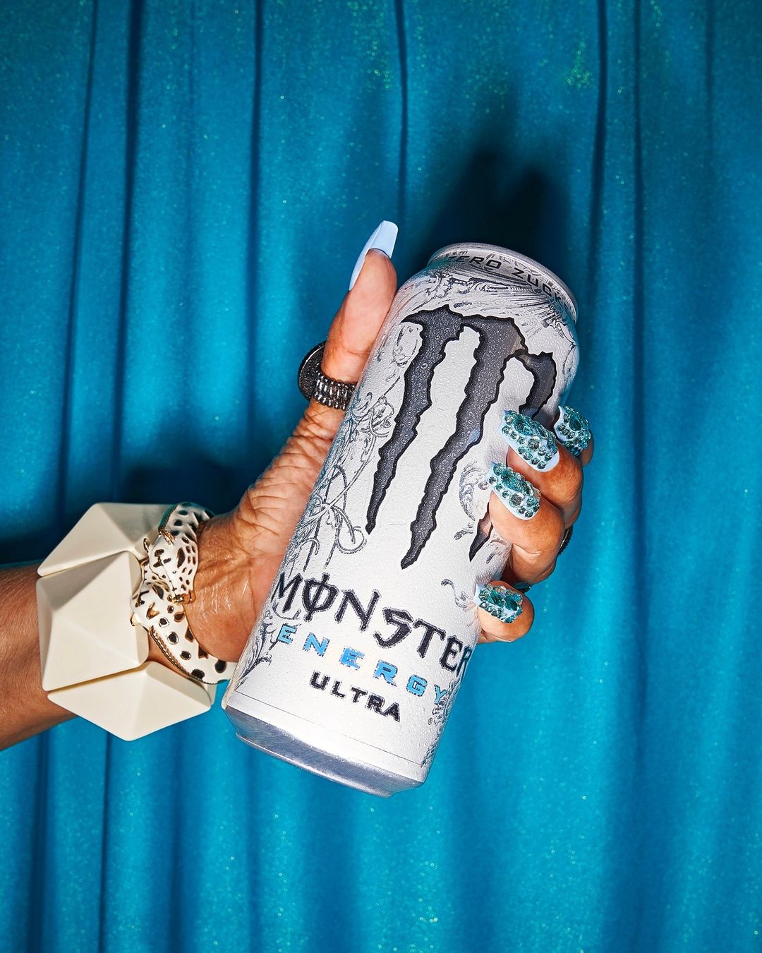 Hand holding a white Monster Energy Ultra can against a turquoise curtain backdrop, accented with statement jewelry and decorative nails.