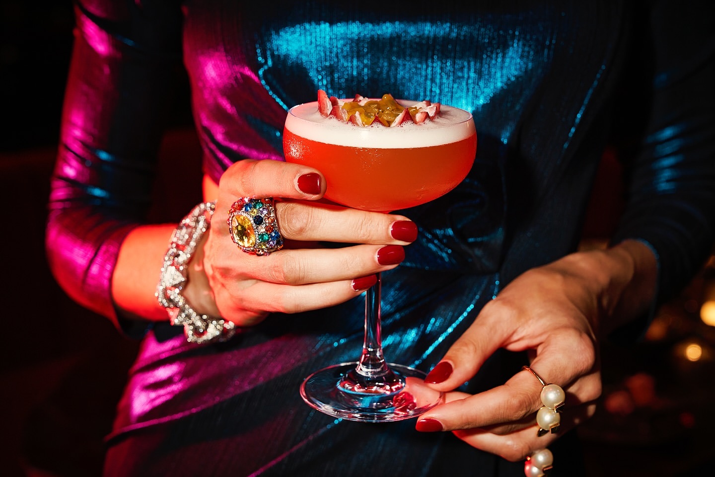 Woman holding a red cocktail garnished with fruit, showcasing an elaborate gemstone ring and pearl ring in a dimly lit setting.