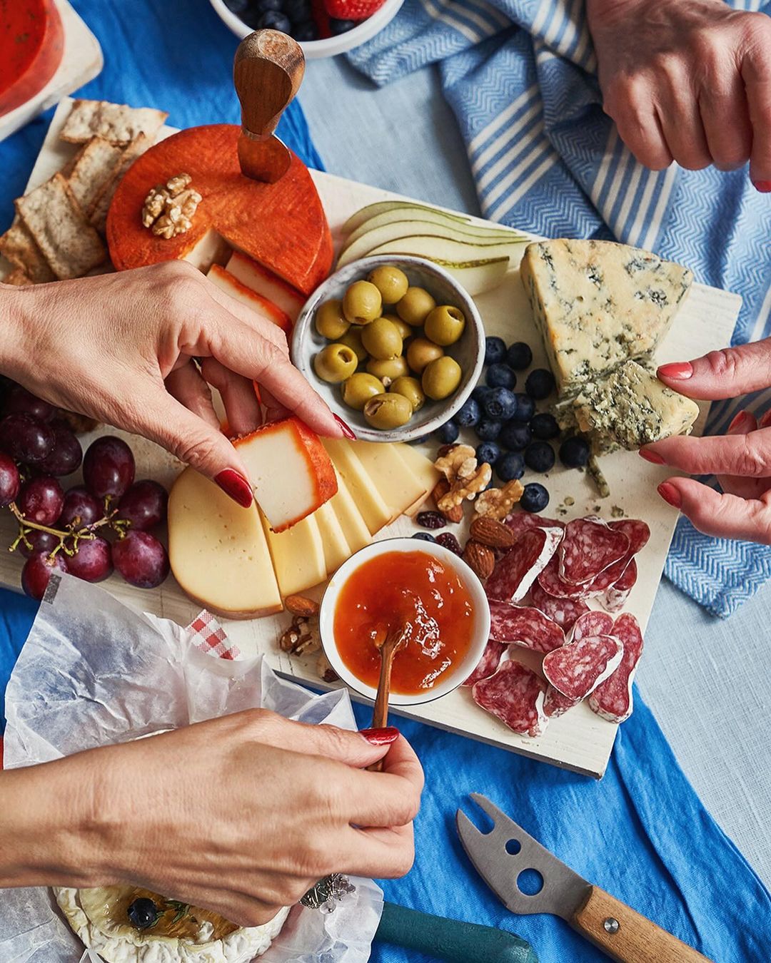 Overhead shot of a cheese board with assorted cheeses, meats, fruits, and nuts, being arranged by two sets of hands on a blue tablecloth.