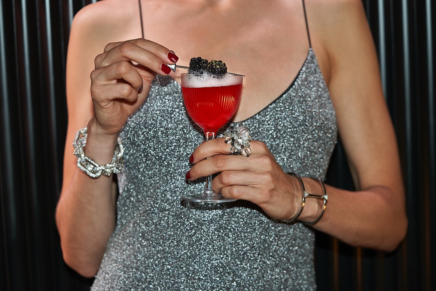 Woman in shimmering dress holds a red cocktail with blackberries, posing in a dark, sleek setting with jewelry accents.