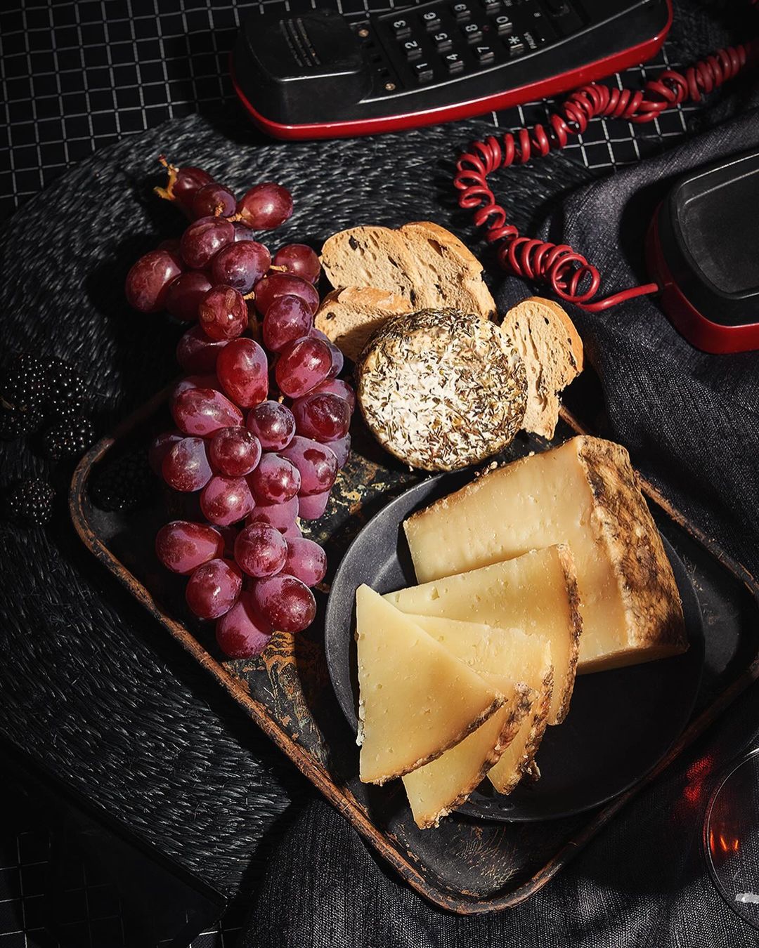 An overhead shot captures a delectable arrangement of cheese, grapes, and bread on a tray, complemented by a vintage telephone and dark textiles.