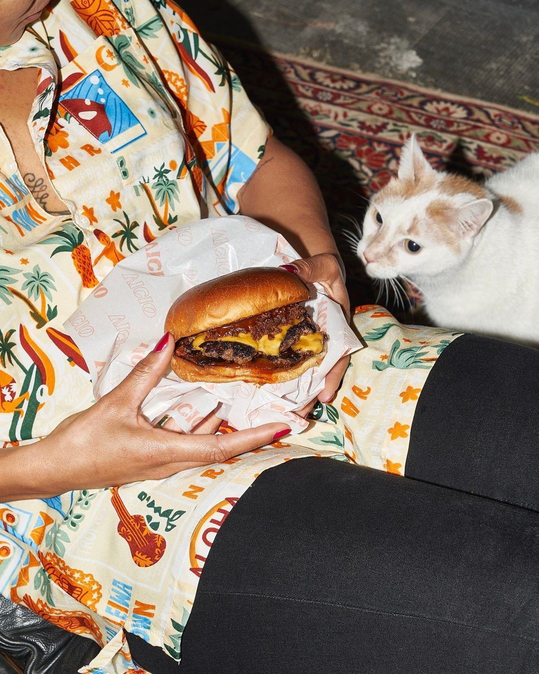 Woman holds a burger while a cat looks on. The burger is wrapped in branded paper and the woman wears a Hawaiian shirt.