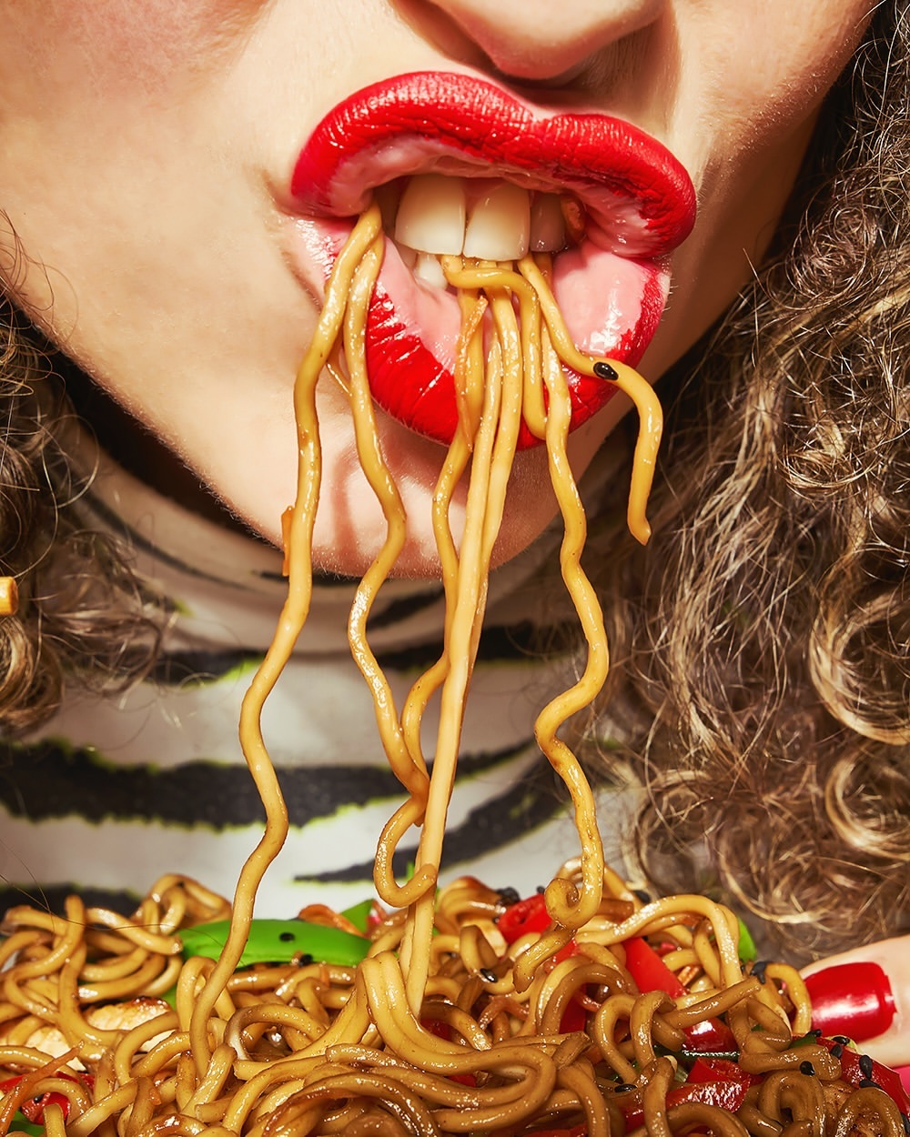 Close-up of person eating noodles with bright red lipstick, showing curly hair, striped top, and focus on the mouth and noodle action.