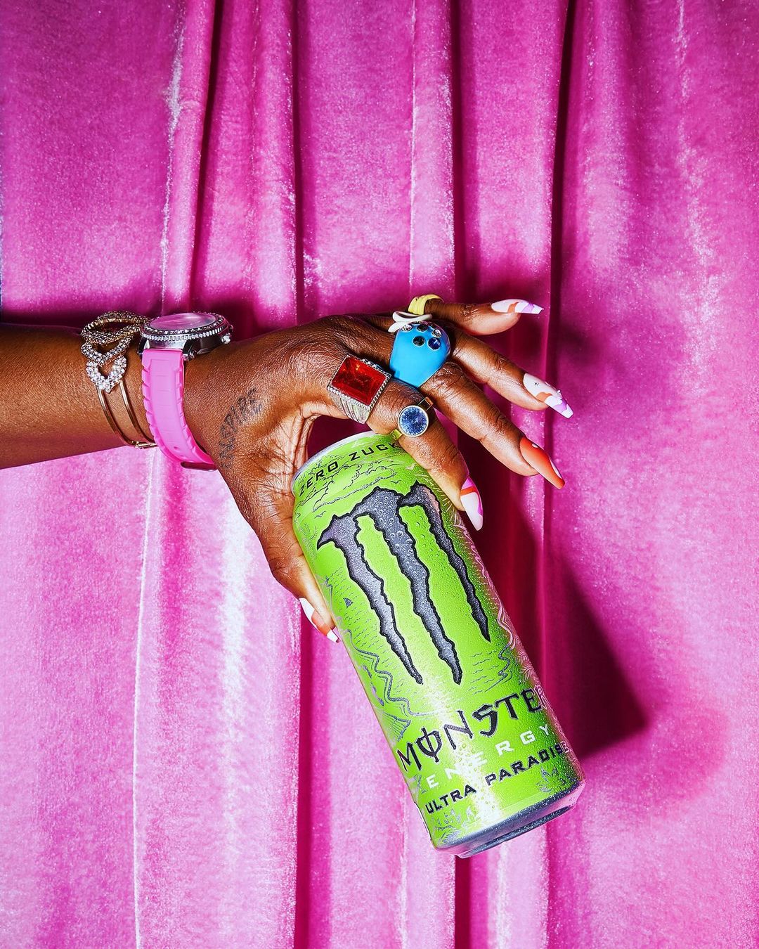Close-up of a dark-skinned hand holding a green Monster Energy drink against a pink curtain background with jewelry on fingers and wrist.