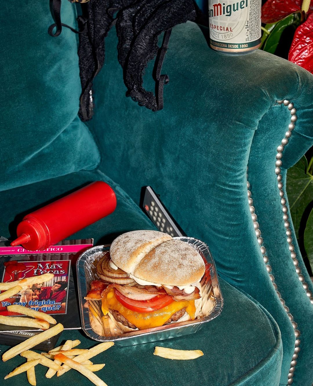 Overhead shot of cheeseburger in foil tray, fries, ketchup, beer, black lingerie, and TV remote on a green tufted couch.