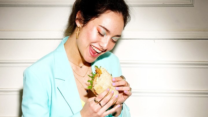 Smiling woman in aqua blazer enjoys a sandwich indoors. Her colorful makeup and gold jewelry add a trendy touch.