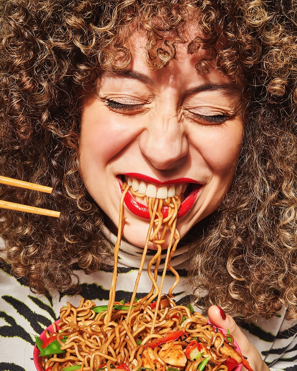Woman with curly hair eating noodles with chopsticks from a pink bowl; she wears a black and white zebra-striped shirt.