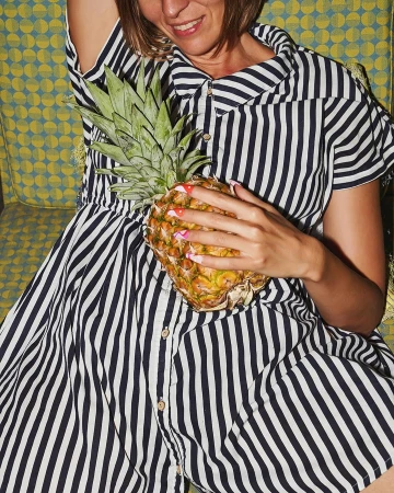 Woman in a navy and white striped dress holding a fresh pineapple against a patterned yellow chair