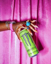 Close-up of a person holding a green energy drink can against a draped pink velvet curtain background.