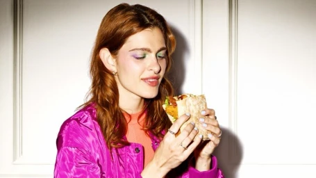 Close-up portrait of a red-haired woman wearing a vibrant magenta jacket holding and eating a filled sandwich
