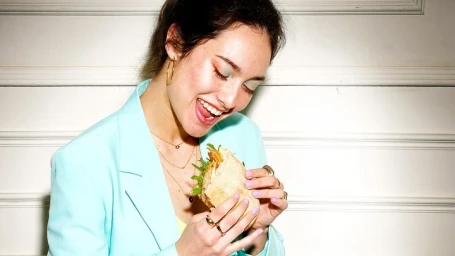 Smiling woman in light blue blazer holding and eating a sandwich against white paneling