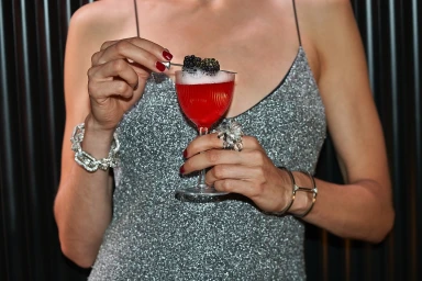 Woman in a silver sparkly dress holding a red cocktail garnished with blackberries against a dark ribbed background