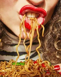 Close-up of a person with bright red lipstick eating noodles from a bowl held near the mouth, featuring curly hair and patterned top.