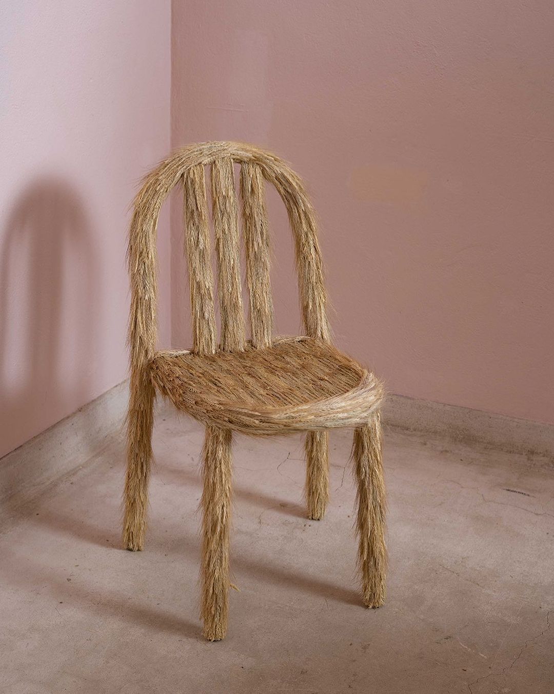 An artistic chair made entirely of straw, set against a muted pink wall, creating a visually striking and minimalist furniture portrait.