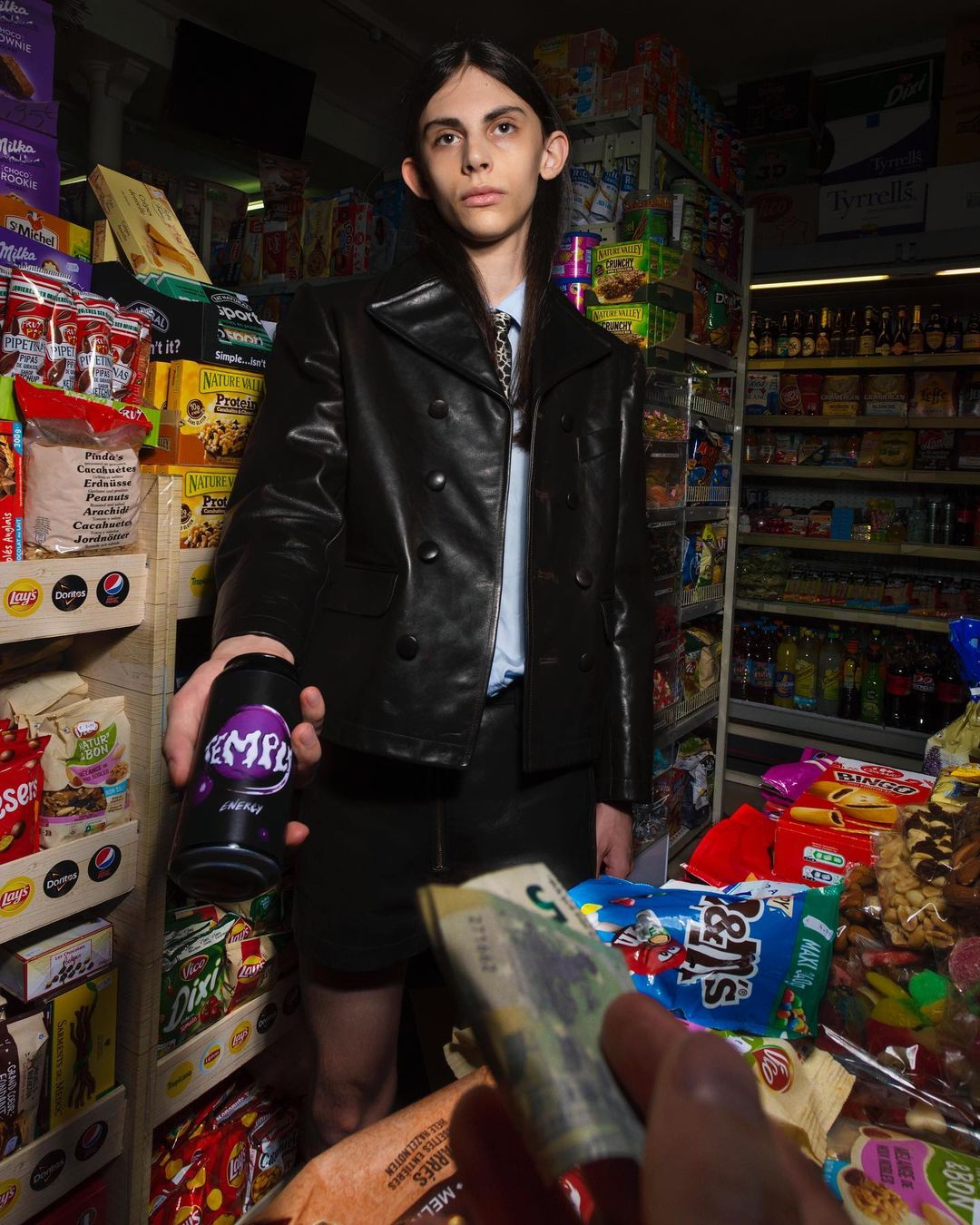 Young adult in black leather jacket offers black can with cash in a convenience store setting with shelves stocked with groceries.