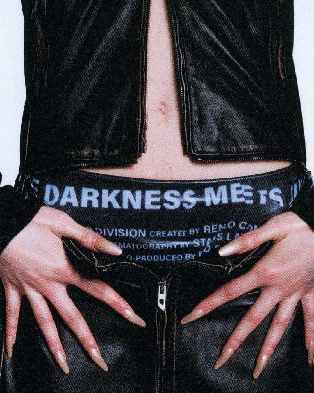 Close-up fashion shot of a woman with long nails wearing a black leather jacket and pants with printed waistband, posing indoors.