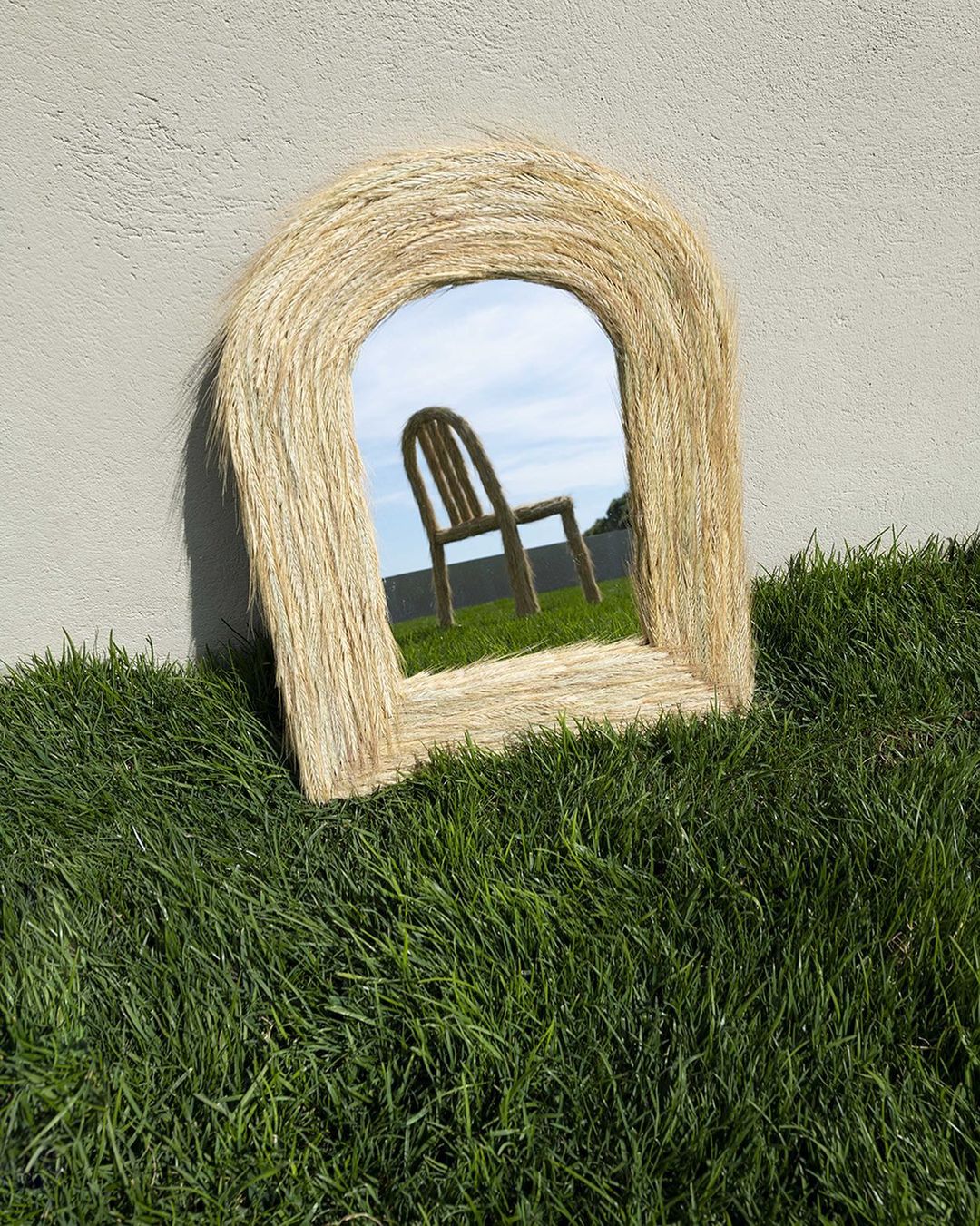 Mirror with straw frame reflecting a chair in a green lawn against a stucco wall, creating a surreal and outdoor decorative accent.